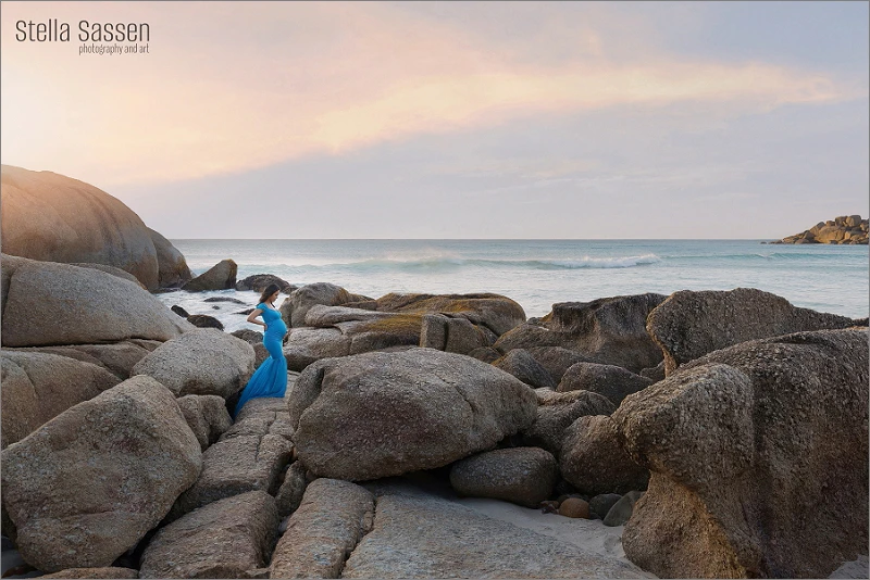 A beautiful maternity shoot at a Cape Town beach during sunset. The expectant mother in a flowing blue dress stands on the rocks, enjoying the stunning view as the sun sets over the ocean